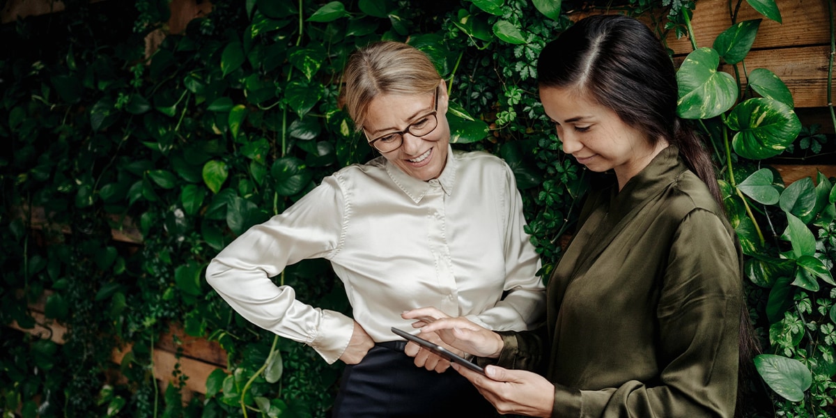 Two women look at tablet in front of leafy background