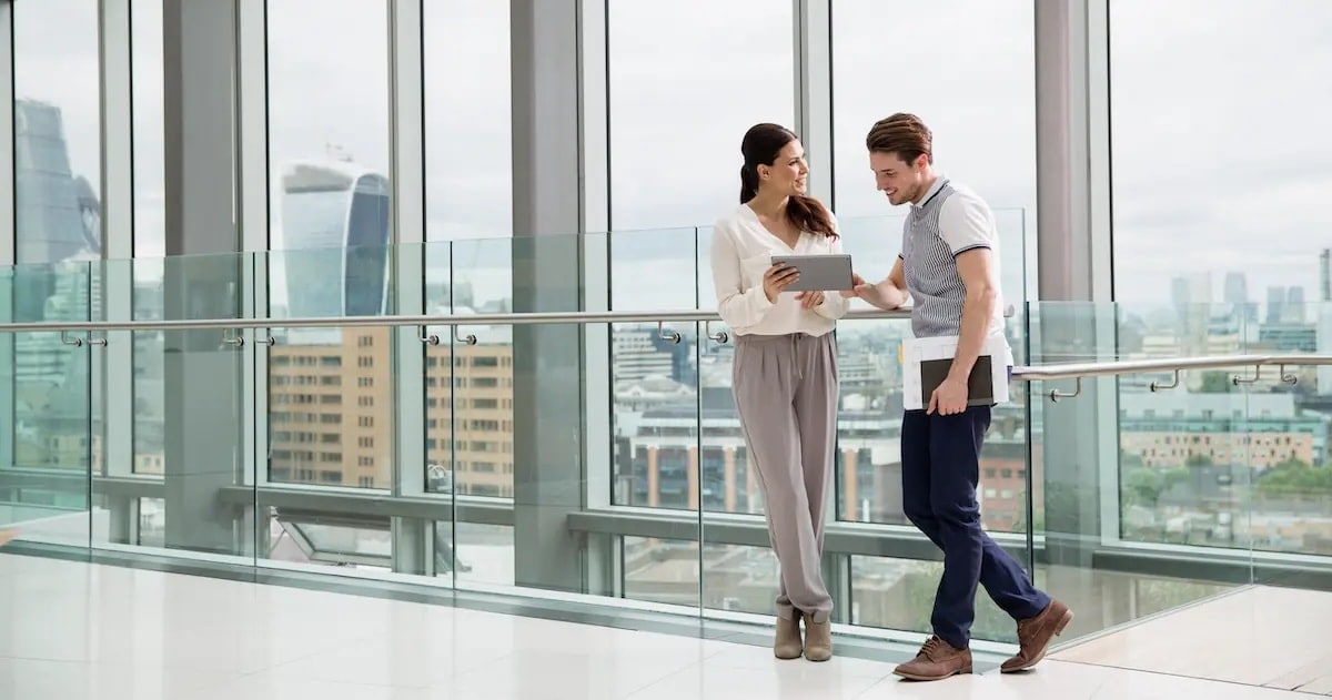 TwA man and a woman standing in front of a glass window in an office looking at a tablet