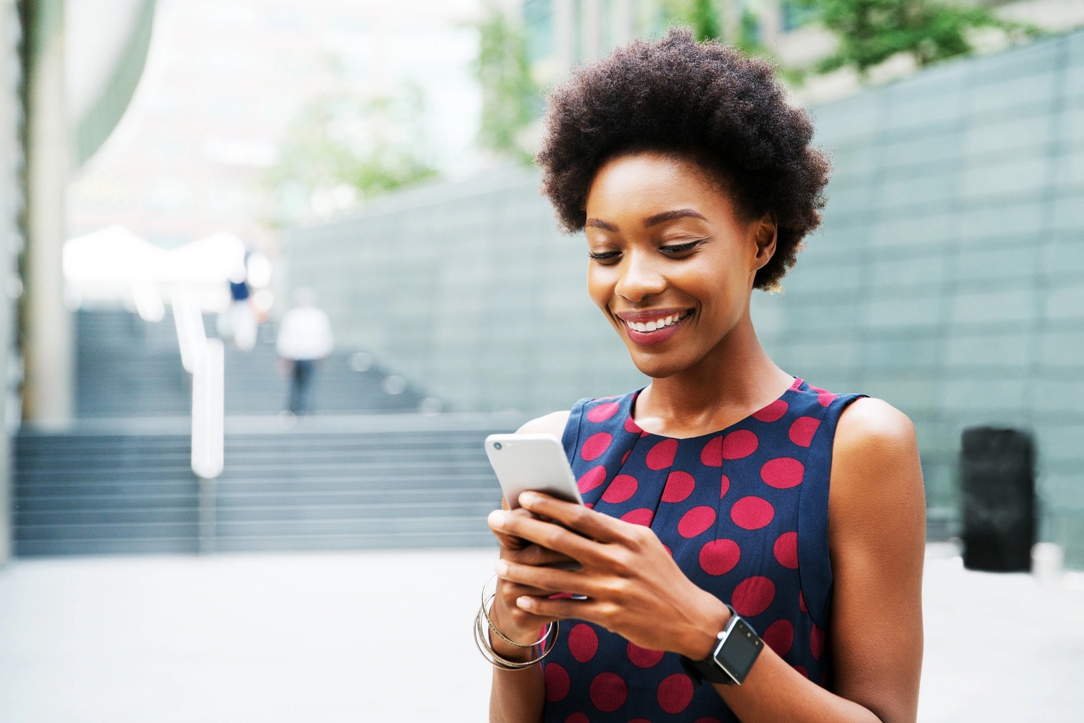 Woman in polka-dot shirt smiles at phone while standing outdoors
