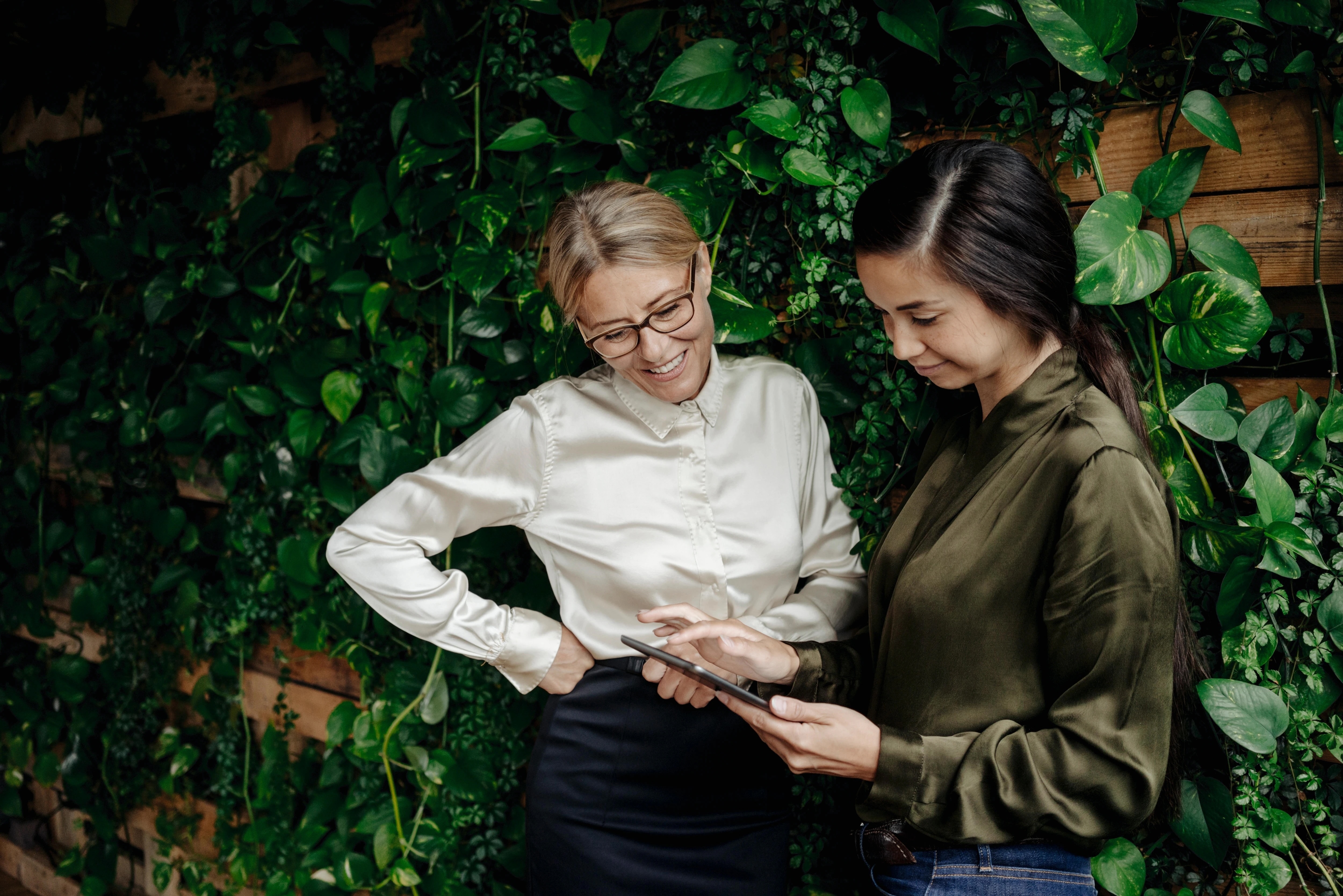 Two women look at tablet in front of leafy background