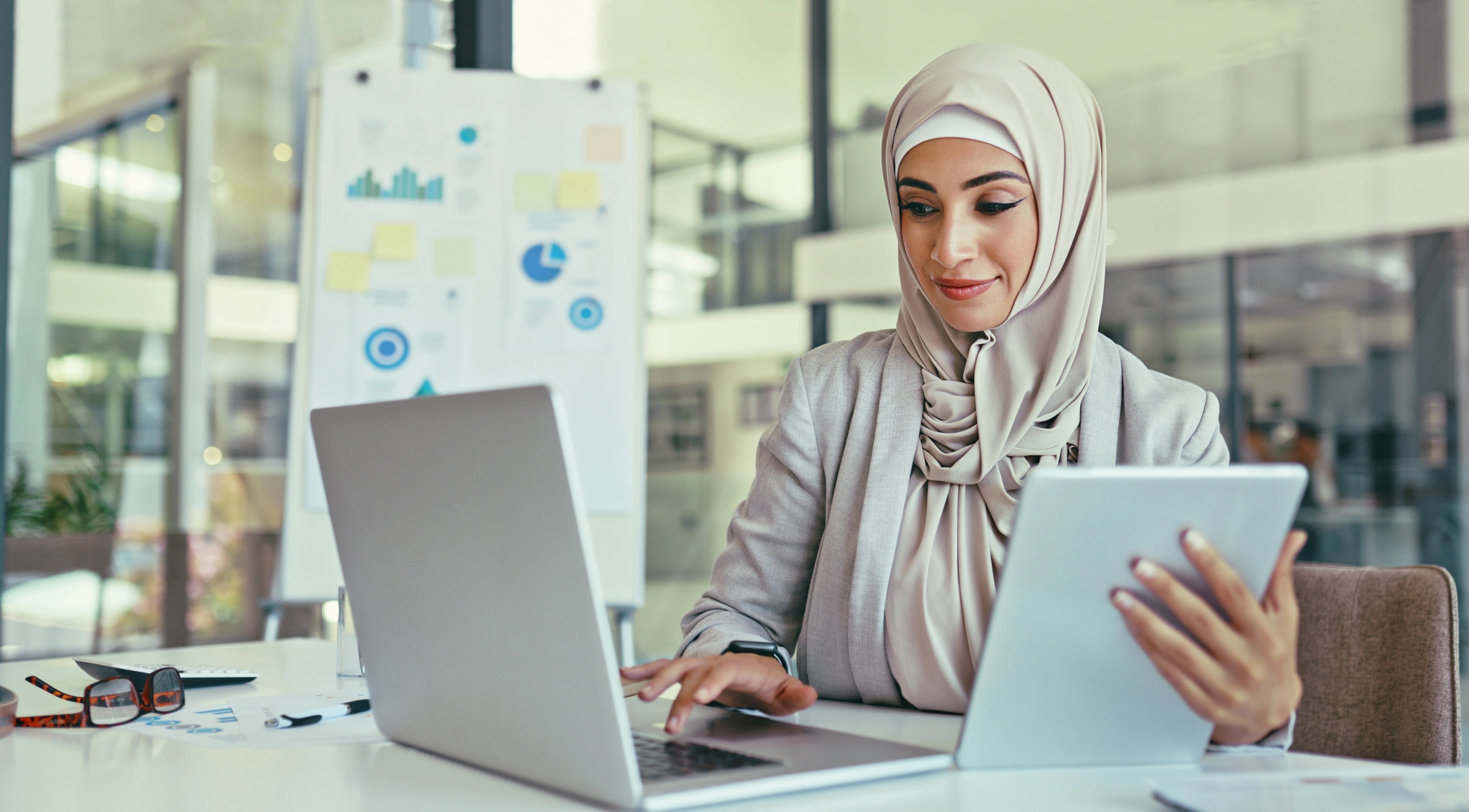 Woman wearing hijab multitasks on tablet and laptop