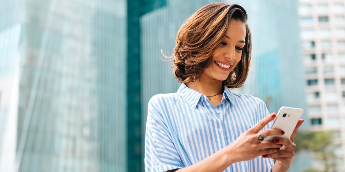 Woman with short brown hair stands outside of a building looking down at her phone 